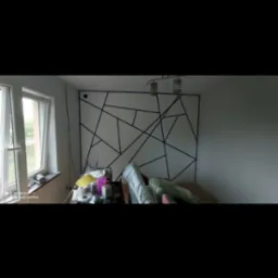 Przygotowanie ściany do malowania z geometrycznym wzorem wyklejonym taśmą malarską, widoczne narzędzia i materiały malarskie na stole w pokoju.