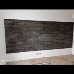 Ściana wykończona dekoracyjnym kamieniem w odcieniach szarości i brązu, widoczne gniazdka elektryczne, podłoga zabezpieczona kartonem podczas prac wykończeniowych.