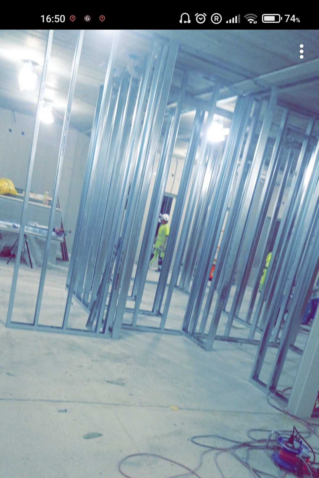 Metalowa konstrukcja szkieletowa ścian działowych w trakcie montażu, widoczni pracownicy w odblaskowych ubraniach, przewody elektryczne na podłodze.