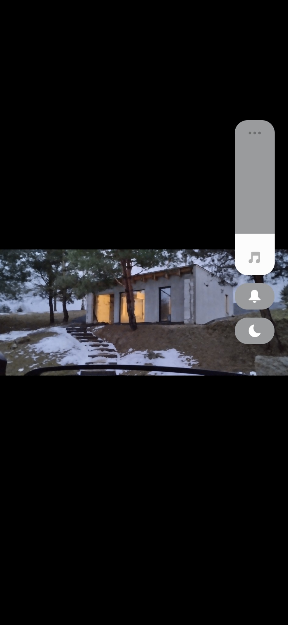 Niedokończony budynek w stanie surowym, z oknami bez szyb, otoczony drzewami i resztkami śniegu, widoczny zza szyby samochodu.