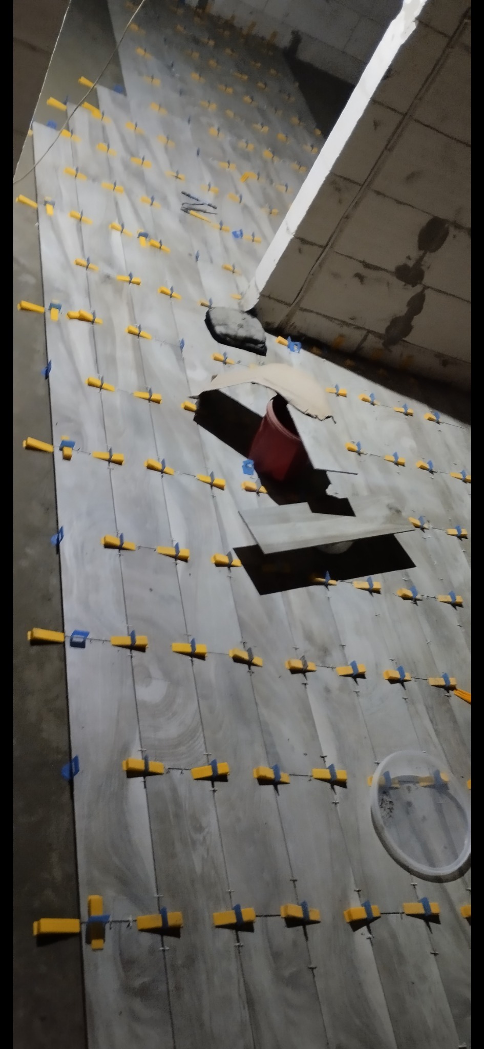 Układanie szarych płytek na ścianie z użyciem żółtych i niebieskich krzyżyków dystansowych, widoczny czerwony pojemnik i wiadro z klejem.