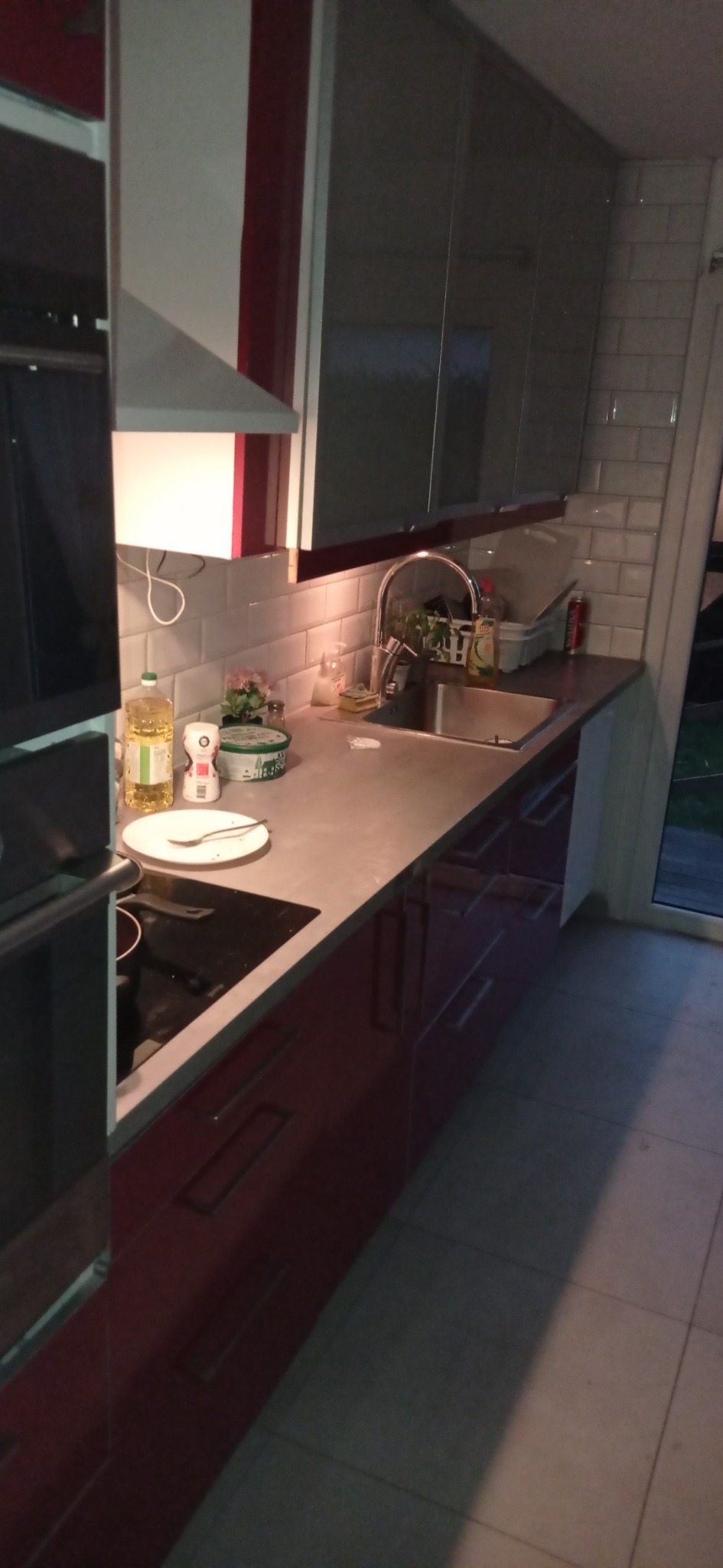 Nowoczesna kuchnia z szarymi szafkami wiszącymi i czerwonymi szafkami dolnymi, biała ściana z płytek, na blacie stoją przyprawy i olej, widoczny zlew i płyta indukcyjna.