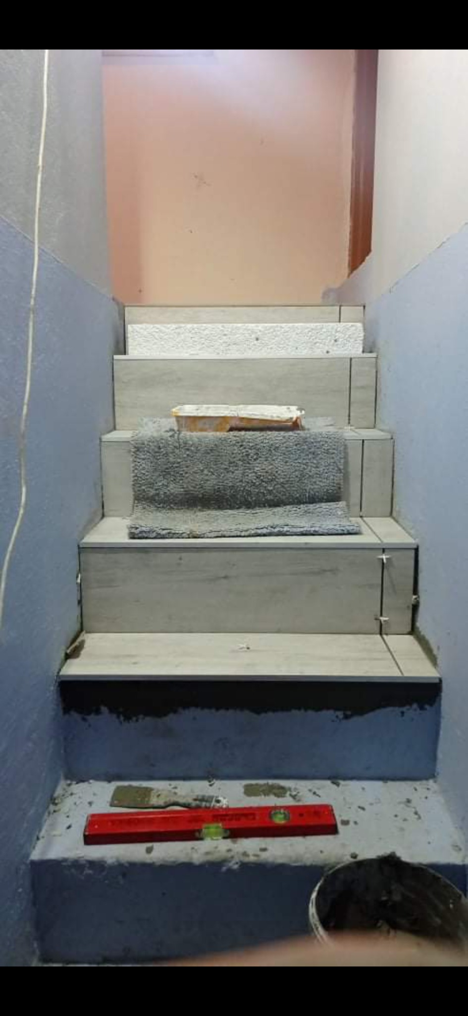 Schody w trakcie układania płytek, widoczny poziomica, szpachelka i wiadro z zaprawą.