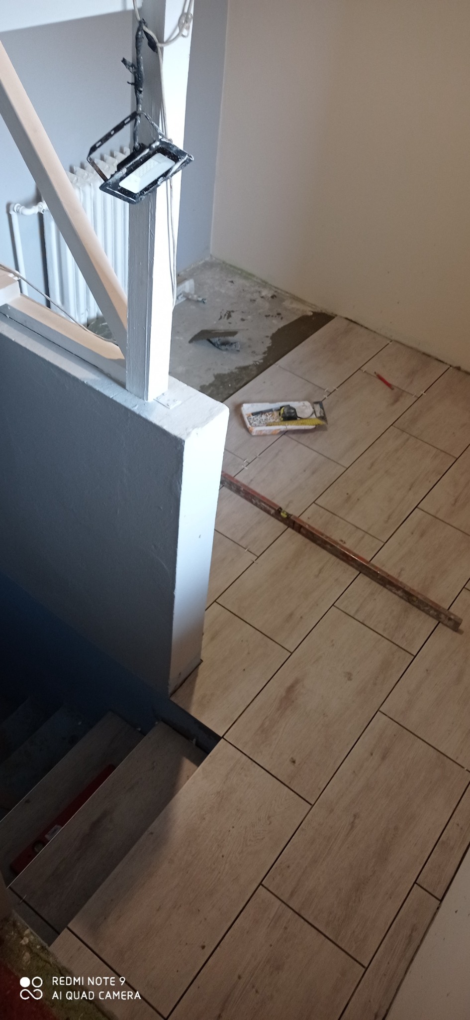 Układanie beżowych płytek podłogowych na schodach i podłodze, widoczne narzędzia glazurnicze, poziomica oraz lampa halogenowa na statywie.