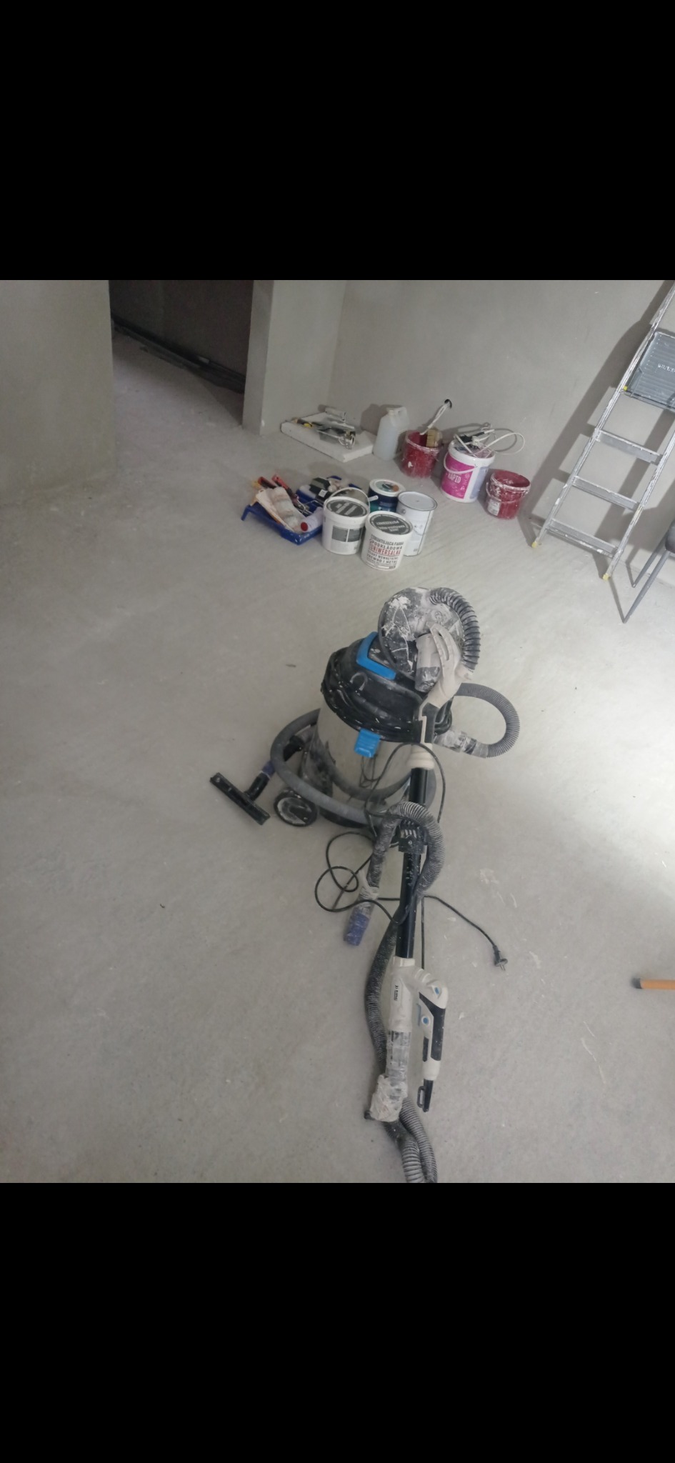 Przygotowania do prac wykończeniowych: szlifierka do gładzi z odkurzaczem przemysłowym, pędzle, wałki malarskie, drabina i puszki z farbami w surowym pomieszczeniu.