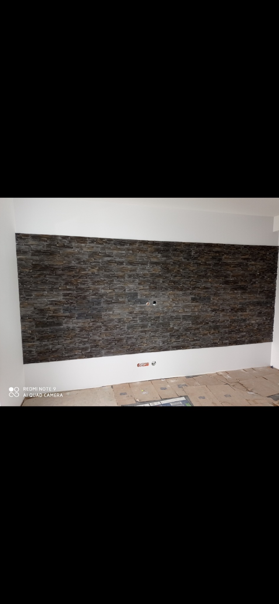 Ściana wykończona dekoracyjnym kamieniem w odcieniach szarości i brązu, widoczne gniazdka elektryczne, podłoga zabezpieczona kartonem podczas prac wykończeniowych.