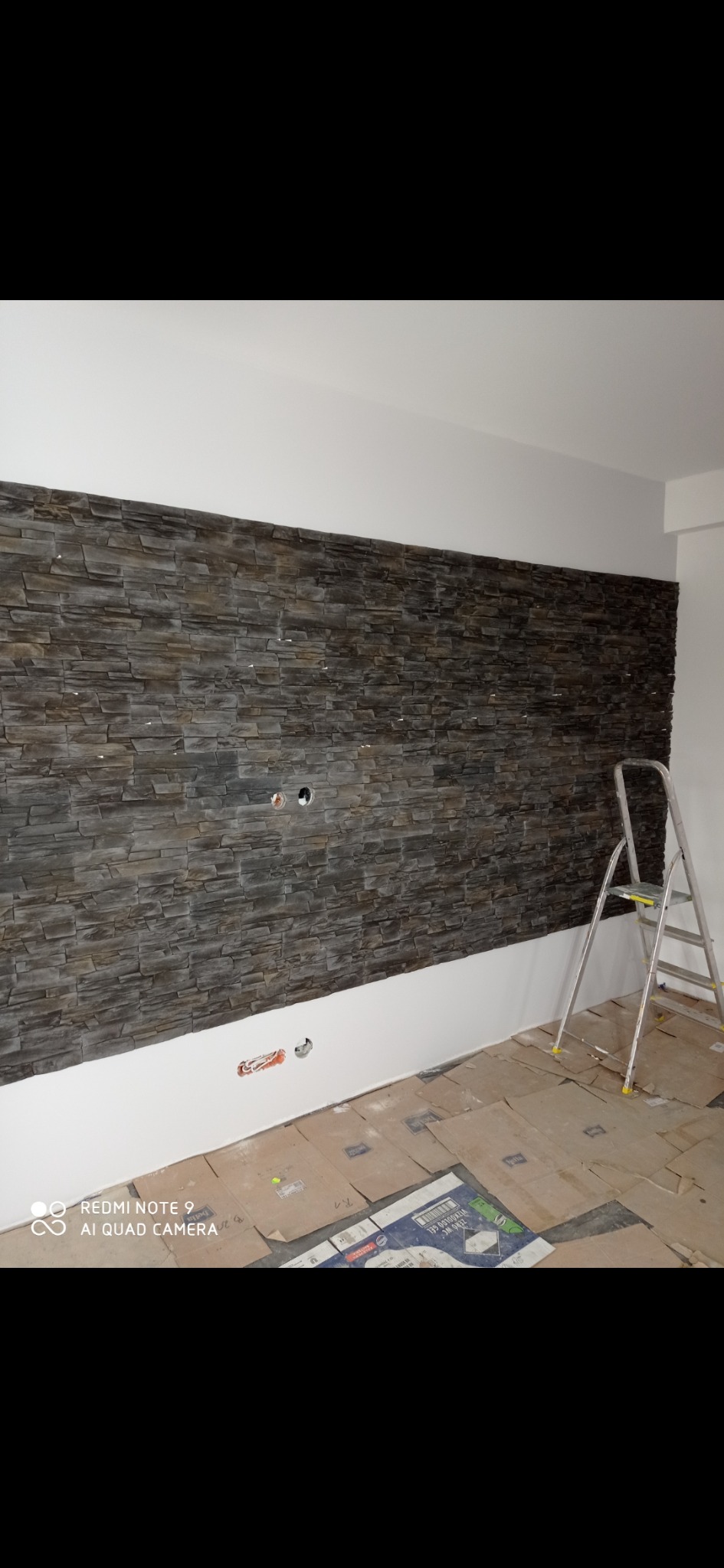 Ściana wyłożona dekoracyjnym kamieniem w odcieniach szarości i brązu, z widocznymi puszkami elektrycznymi, zabezpieczona podłoga kartonem, drabina oparta o ścianę.