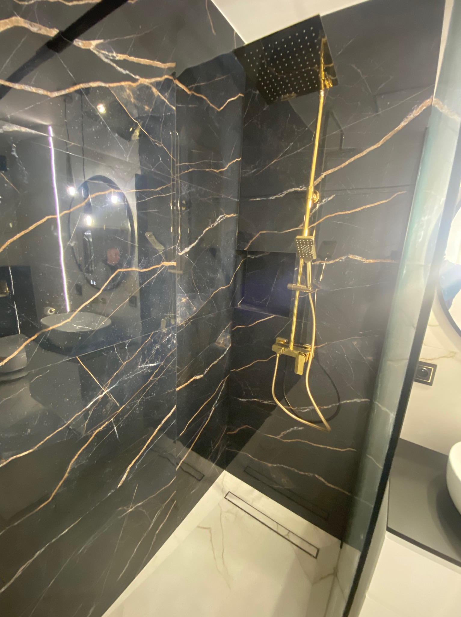 Kabina prysznicowa wyłożona czarnym marmurem z złotymi żyłami, złota armatura, w tle widoczne odbicie umywalki i lustra.