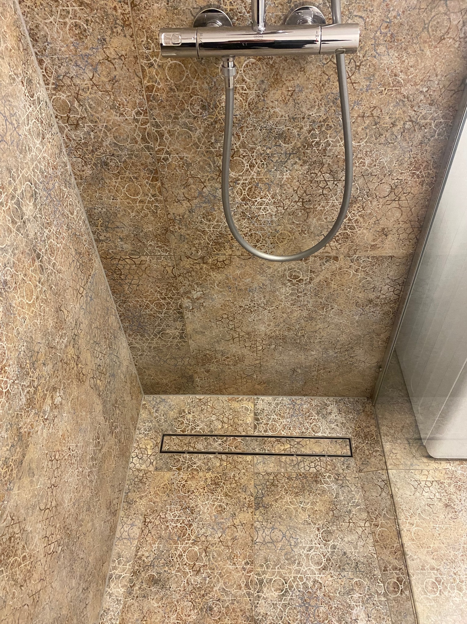 Kabina prysznicowa wykończona brązowymi płytkami z geometrycznym wzorem, widoczny odpływ liniowy i bateria prysznicowa Grohe.