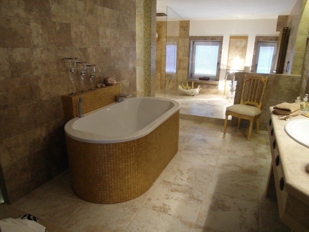 Luksusowa łazienka z wanną obłożoną mozaiką w odcieniach złota, marmurowa podłoga i ściany, szklana kabina prysznicowa w tle, eleganckie krzesło i zestaw szklanek na półce.