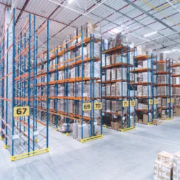 Wysokie regały magazynowe z numeracją, wypełnione zapakowanymi towarami, widok z g&oacute;ry na posadzkę z paletami gotowymi do załadunku.