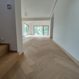 Jasny parkiet ułożony we wzór jodełki francuskiej w przestronnym, nowym wnętrzu z dużymi oknami i drewnianymi schodami.