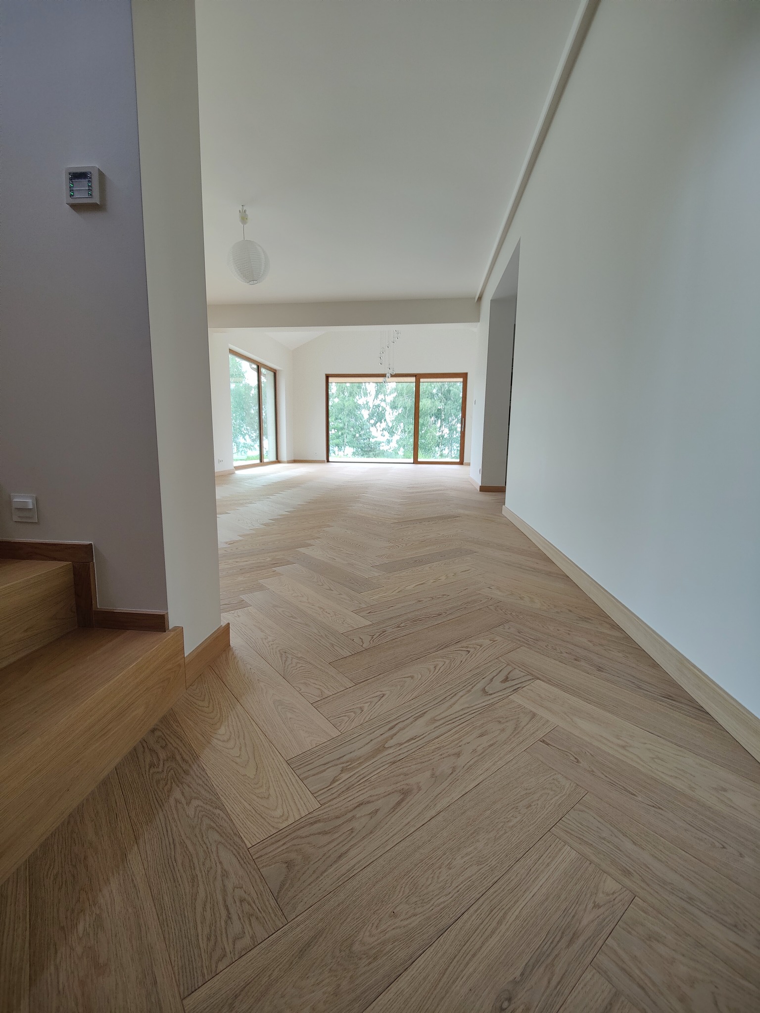 Jasny parkiet ułożony we wzór jodełki francuskiej w przestronnym, nowym wnętrzu z dużymi oknami i drewnianymi schodami.