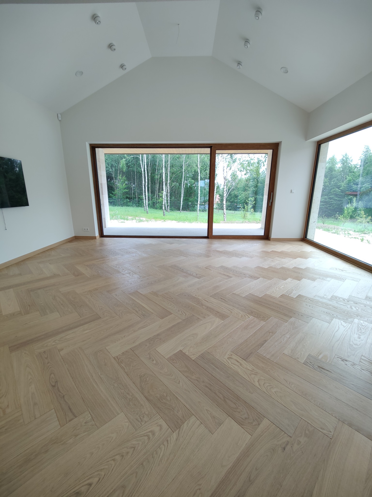 Jasny parkiet ułożony we wzór jodełki francuskiej w przestronnym, nowoczesnym salonie z dużymi oknami przesuwnymi wychodzącymi na zielony ogród.