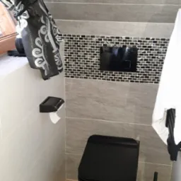 Nowoczesna łazienka z czarną toaletą, mozaiką nad stelażem podtynkowym i drewnopodobnymi płytkami podłogowymi, zasłonięte okno z lewej.