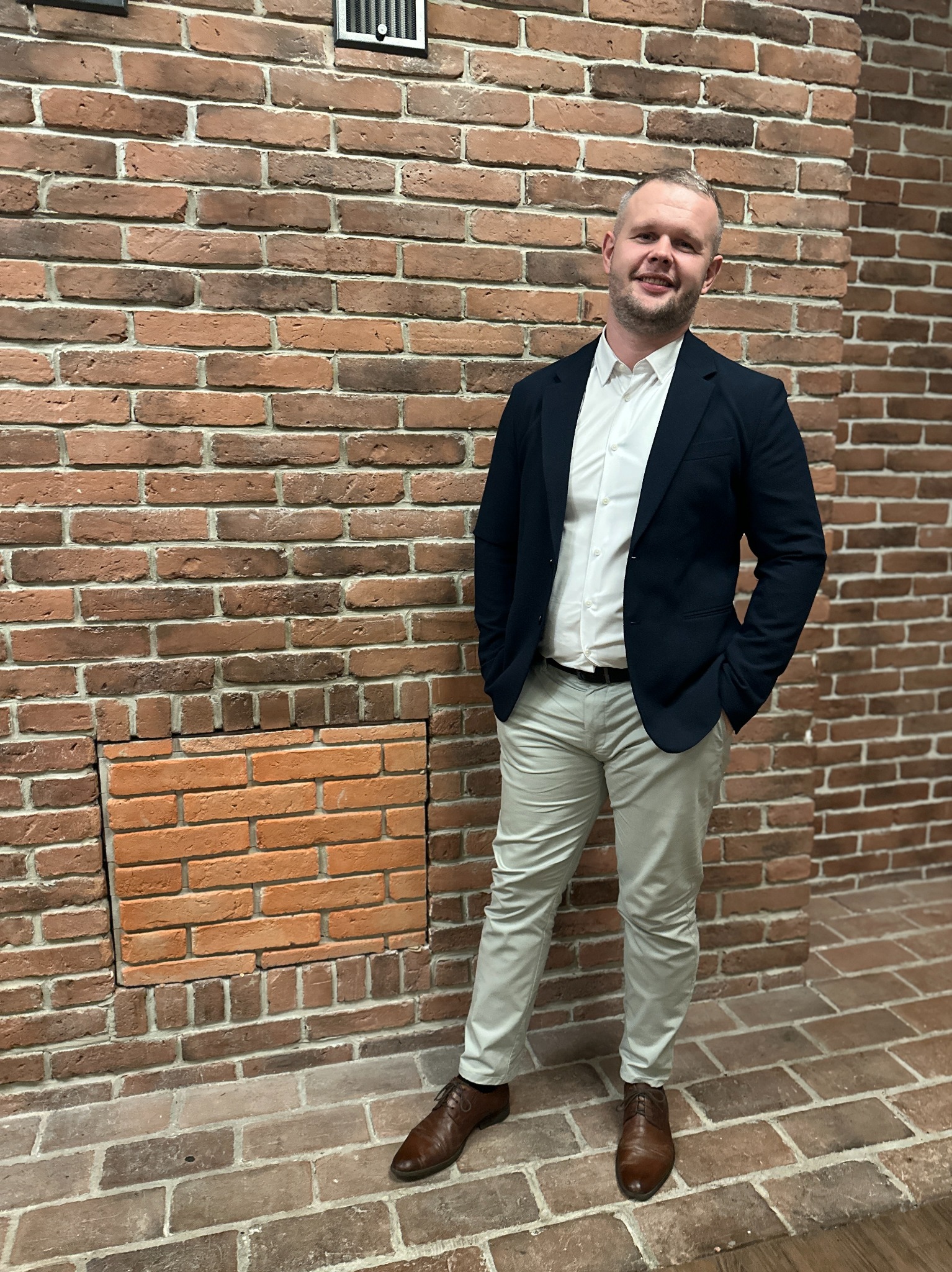 Uśmiechnięty mężczyzna w marynarce i spodniach chinos pozuje na tle ściany z czerwonej cegły z dekoracyjnym wnęką.