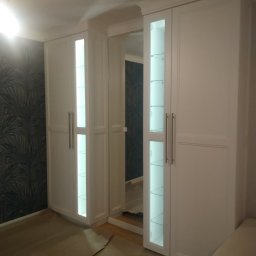 Morti Joanna Sieńkowska - Biała zabudowa meblowa z podświetlanymi witrynami i lustrem w przedpokoju, widoczne zabezpieczenie podłogi kartonem.