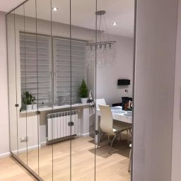 Morti Joanna Sieńkowska - Szafa z lustrzanymi drzwiami przesuwnymi na całej ścianie, odbijająca wnętrze jadalni z żyrandolem kryształowym, roślinami na parapecie i białym stołem.