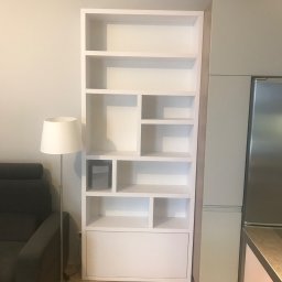 Morti Joanna Sieńkowska - Biały regał wnękowy z asymetrycznym układem półek, wkomponowany w aranżację wnętrza obok zabudowy kuchennej, oświetlony lampą podłogową.