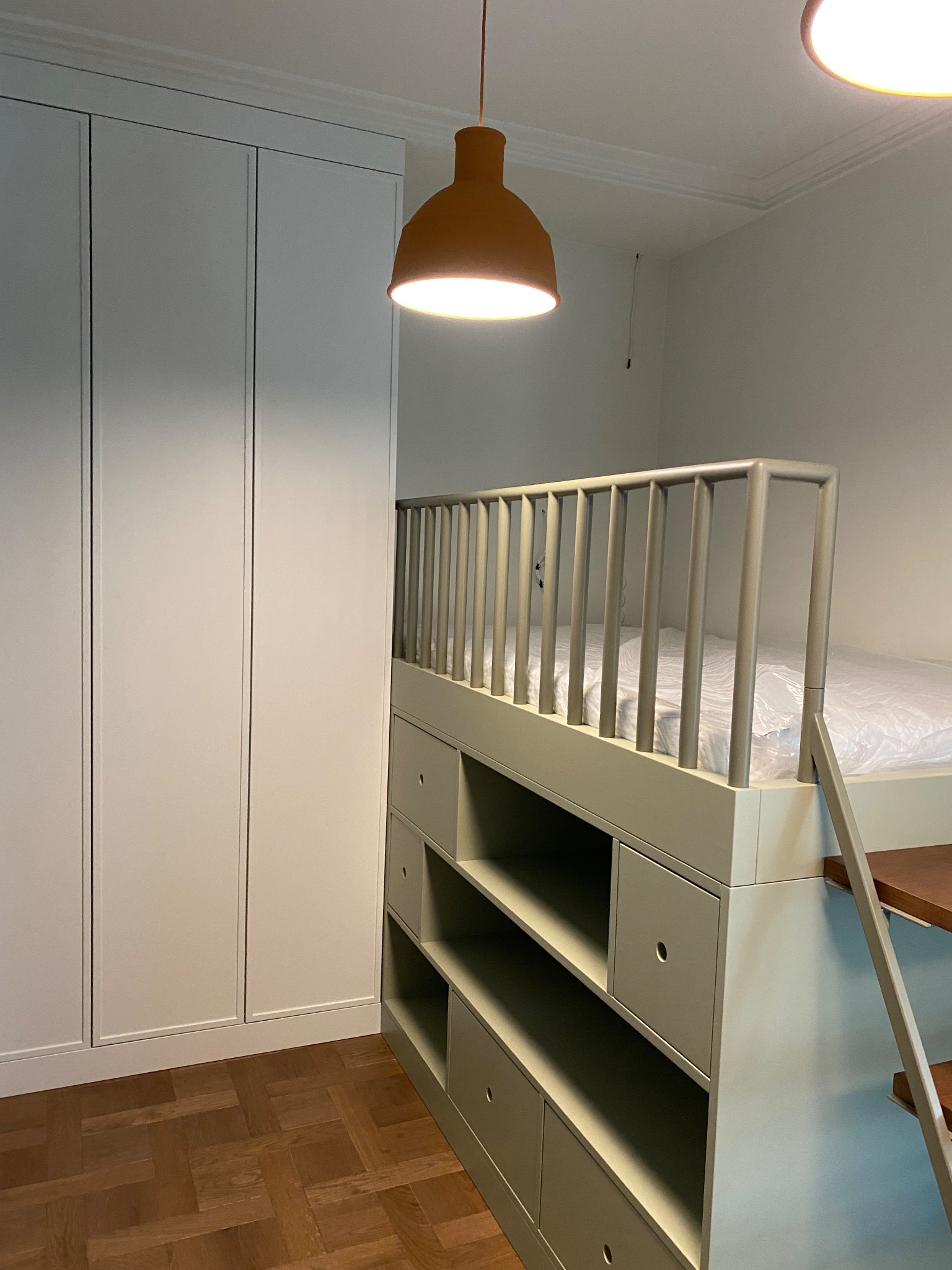 Wysokie łóżko z drabinką, wbudowane w szafę z szufladami i półkami, oświetlone ciepłym światłem lampy wiszącej.