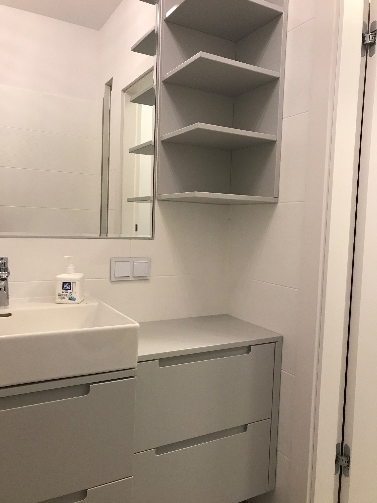 Nowoczesna, szara szafka łazienkowa z umywalką nablatową i otwartymi półkami, białe płytki na ścianie, minimalistyczny styl.