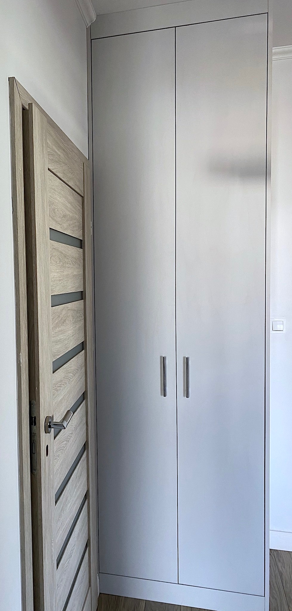 Szara, dwudrzwiowa szafa sięgająca sufitu z podłużnymi, metalowymi uchwytami, widoczna obok otwartych drzwi z drewnopodobnym wykończeniem i ukośnymi, szarymi wstawkami.