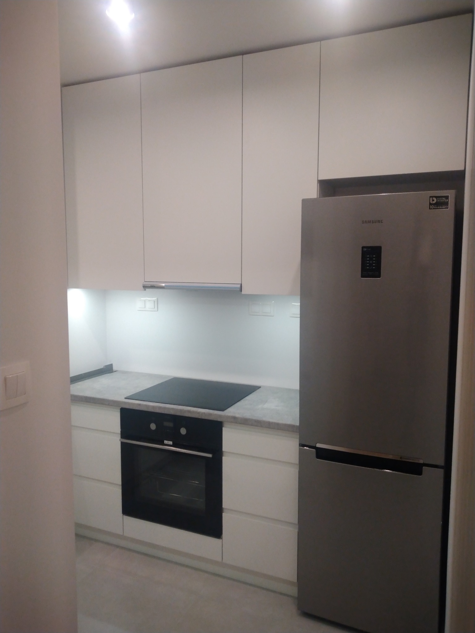 Nowoczesna, minimalistyczna kuchnia z białymi szafkami, szarym blatem i srebrną lodówką Samsung.