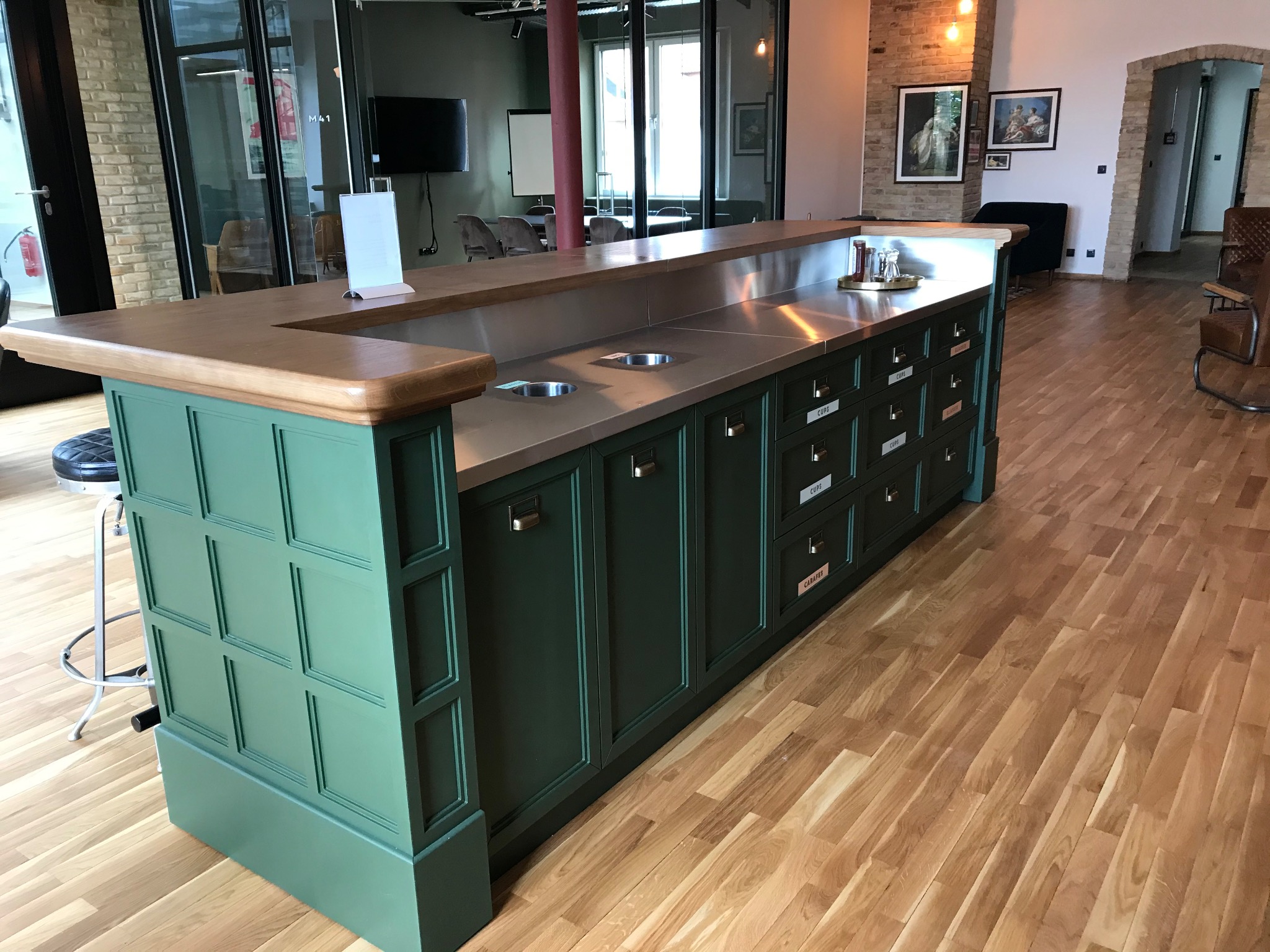 Zielona wyspa kuchenna z drewnianym blatem i metalowym wykończeniem w biurze, z szufladami oznaczonymi napisami, na drewnianej podłodze, w tle biurowe wnętrze.