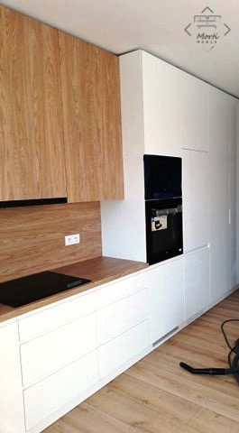 Nowoczesna kuchnia z białymi szafkami i drewnianymi frontami, z wbudowanym piekarnikiem i płytą indukcyjną, widoczny fragment odkurzacza na podłodze.