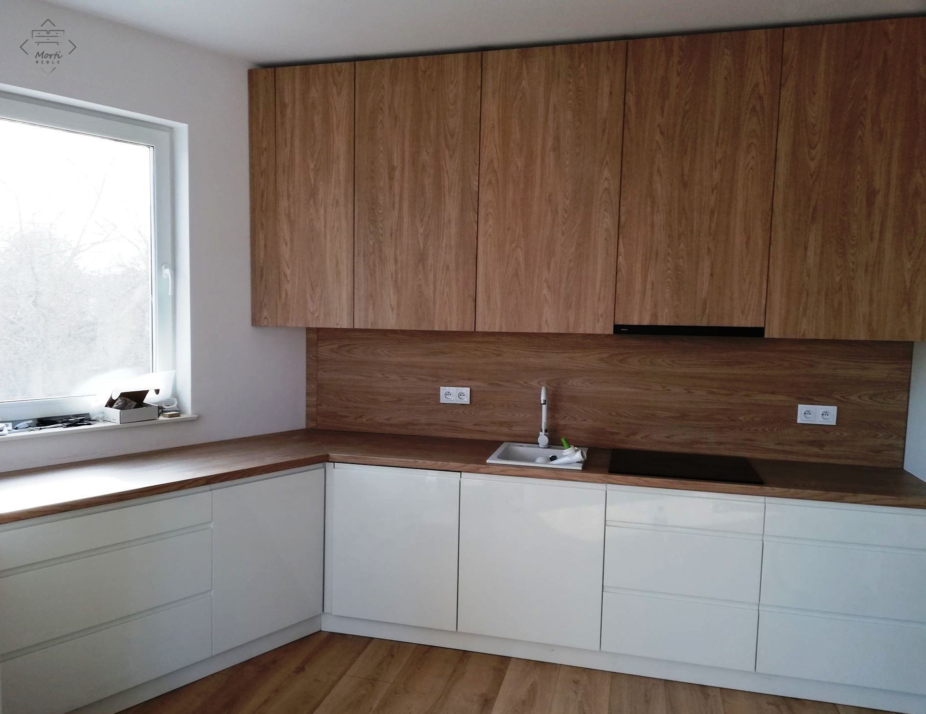 Nowoczesna kuchnia w kształcie litery L z białymi szafkami dolnymi i górnymi szafkami z drewnopodobnym fornirem, drewniany blat, zlewozmywak z baterią, płyta indukcyjna, okno z akcesoriami...