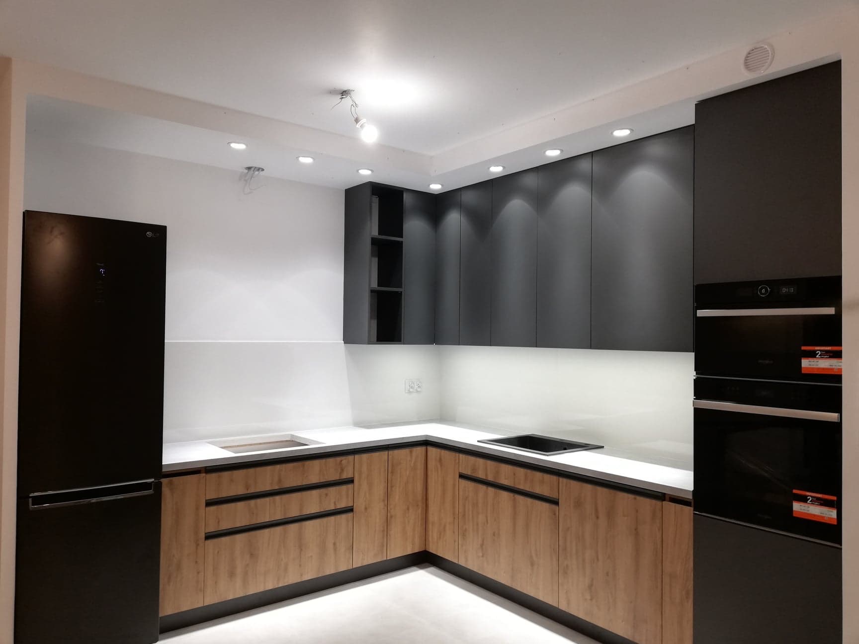 Nowoczesna kuchnia z drewnianymi szafkami dolnymi, czarnymi szafkami górnymi i sprzętem AGD w zabudowie, oświetlona punktowymi lampami sufitowymi.