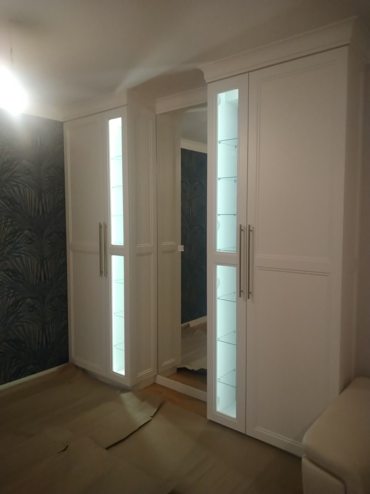 Biała zabudowa meblowa z podświetlanymi witrynami i lustrem w przedpokoju, widoczne zabezpieczenie podłogi kartonem.