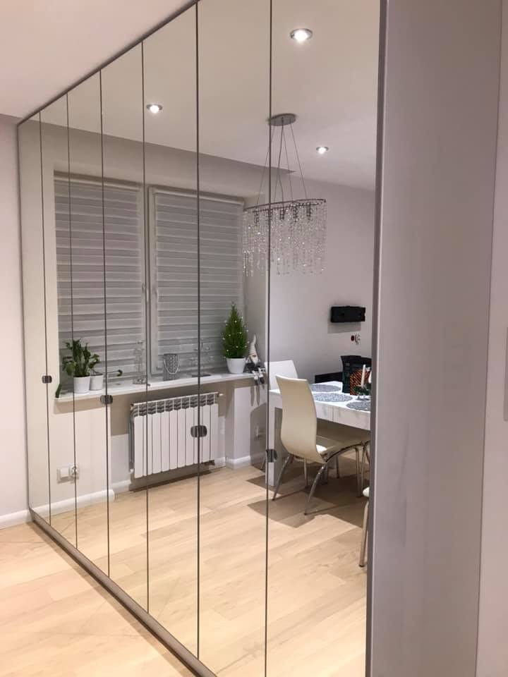 Szafa z lustrzanymi drzwiami przesuwnymi na całej ścianie, odbijająca wnętrze jadalni z żyrandolem kryształowym, roślinami na parapecie i białym stołem.