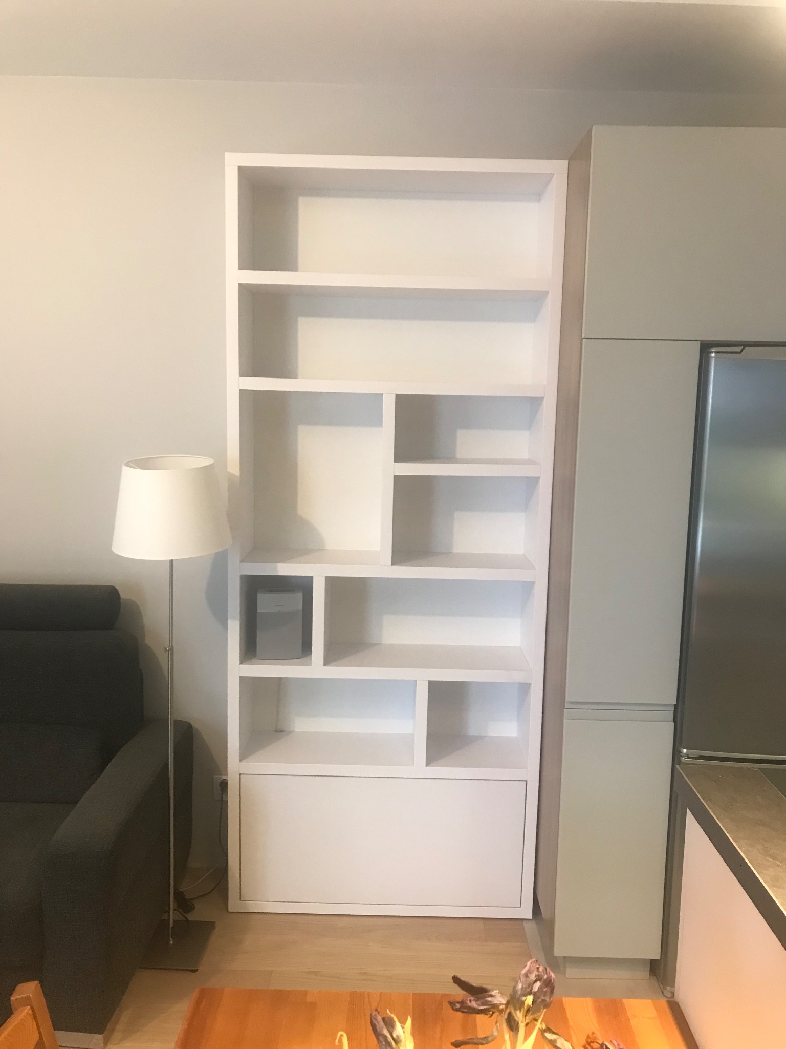 Biały regał wnękowy z asymetrycznym układem półek, wkomponowany w aranżację wnętrza obok zabudowy kuchennej, oświetlony lampą podłogową.