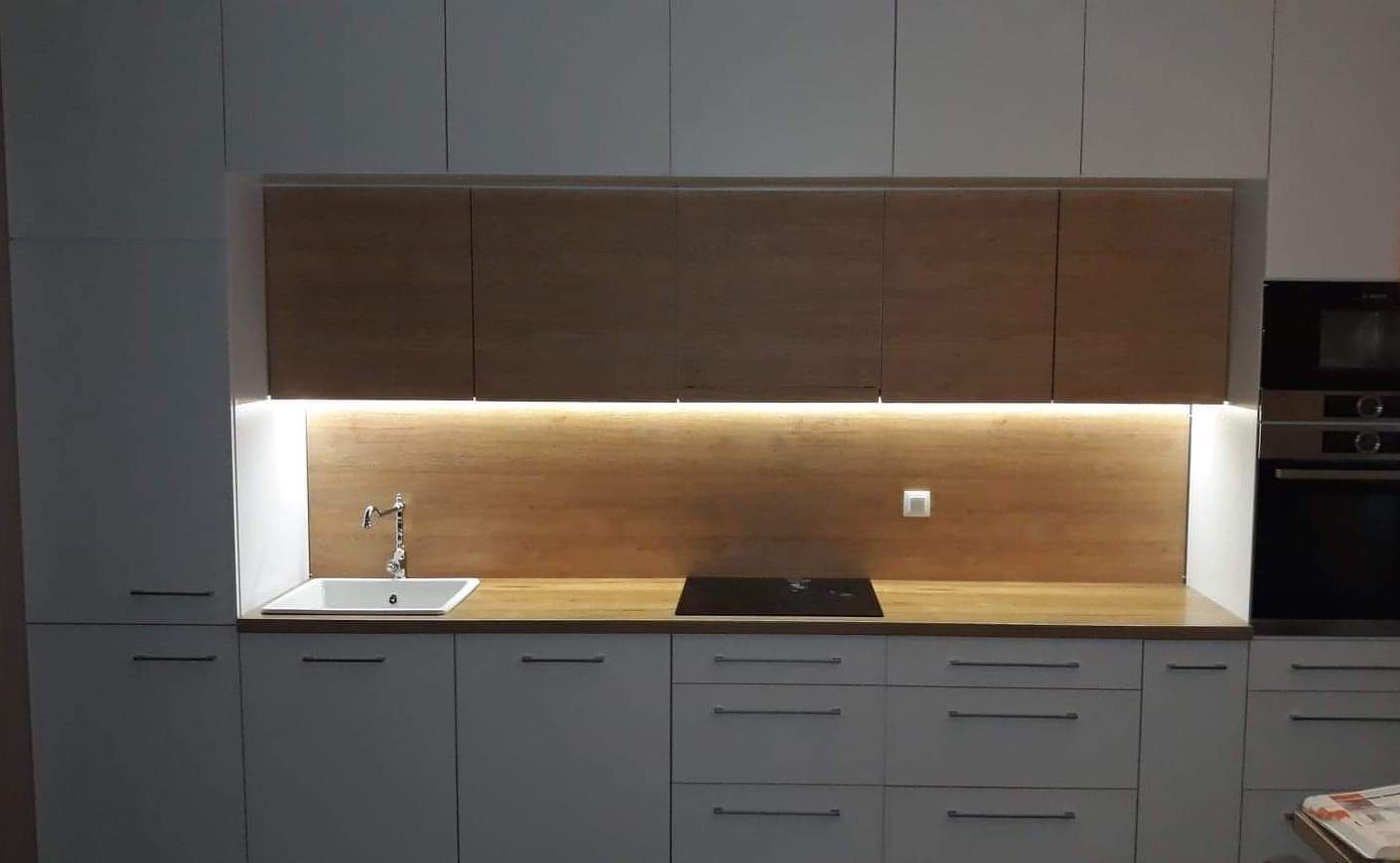 Nowoczesna kuchnia na wymiar z białymi frontami, drewnianym blatem i podświetleniem LED pod szafkami wiszącymi, z widocznym zlewem i płytą indukcyjną.