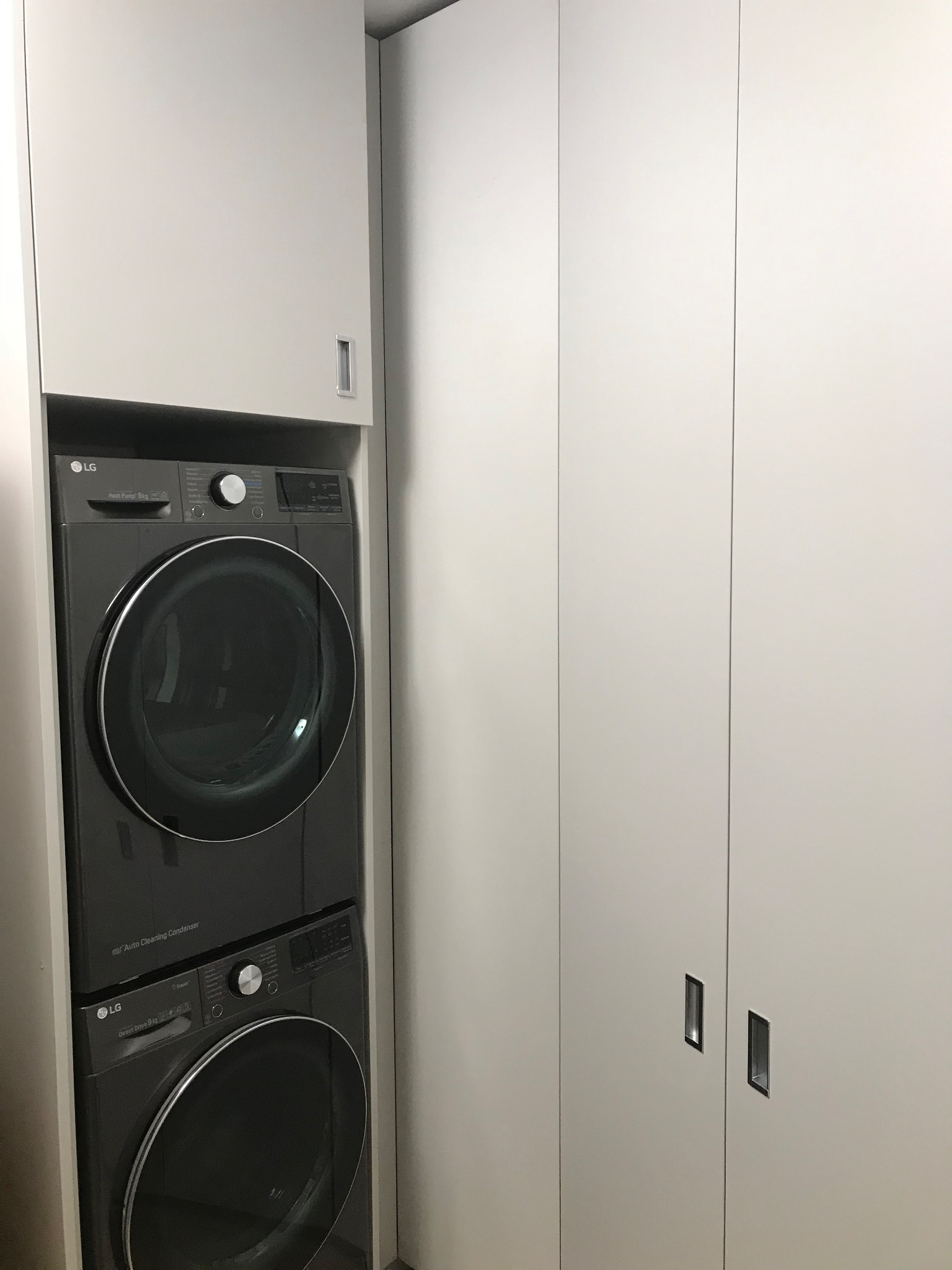 Szara pralka i suszarka LG umieszczone jedna na drugiej w białej szafie wnękowej z minimalistycznymi uchwytami.