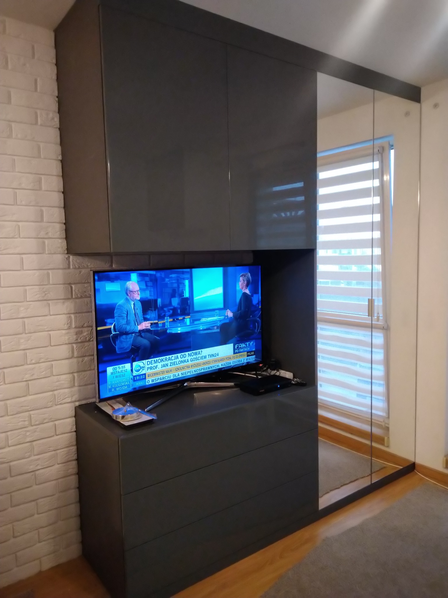 Szara zabudowa meblowa z telewizorem w salonie, widoczna ściana z białej cegły i okno z żaluzjami.