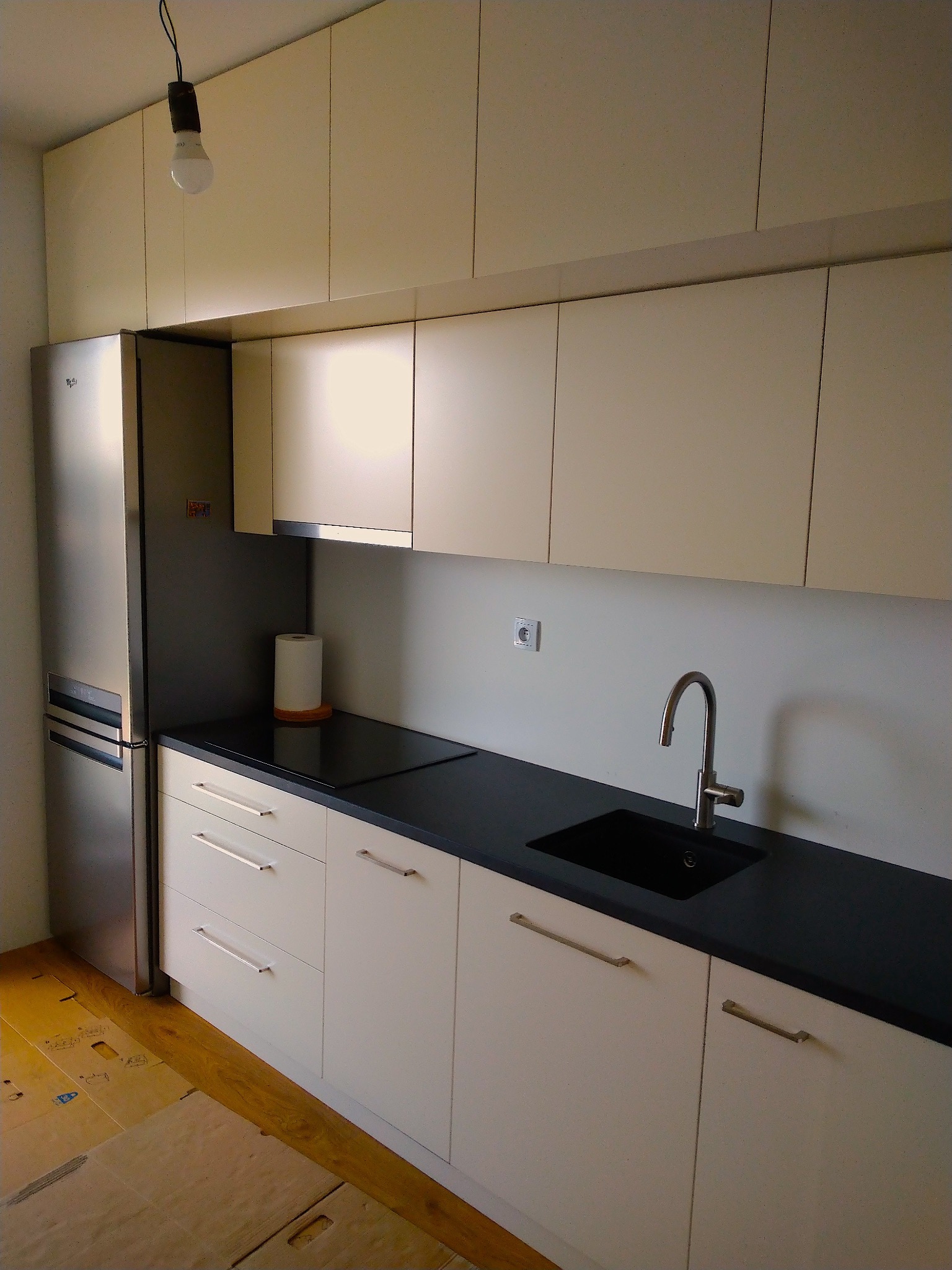 Nowoczesna zabudowa kuchenna z jasnymi szafkami, czarnym blatem i srebrną lodówką; podłoga zabezpieczona kartonem.