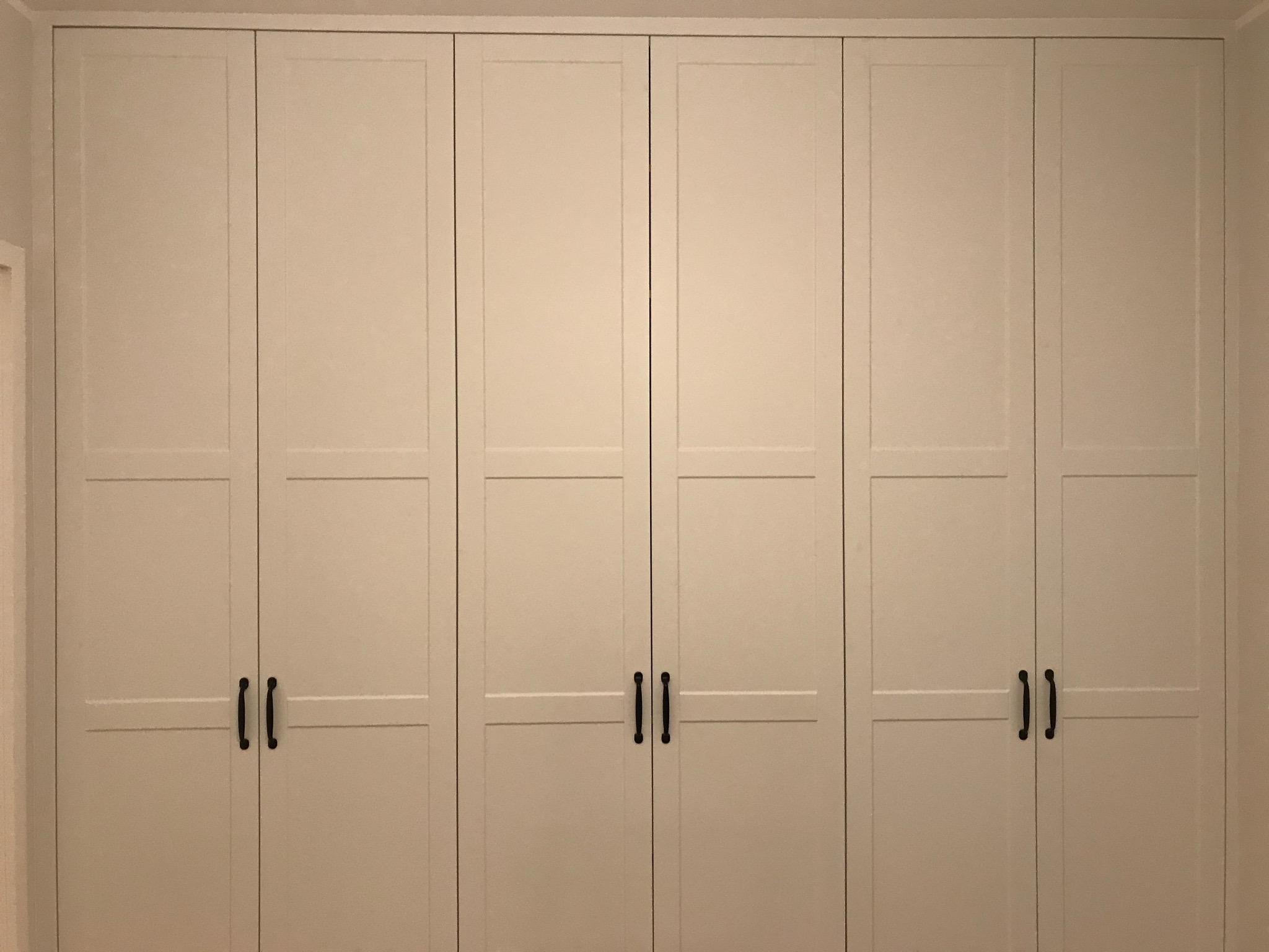 Zamknięta, kremowa szafa wnękowa z sześcioma drzwiami i czarnymi, podłużnymi uchwytami, wypełniająca całą ścianę.