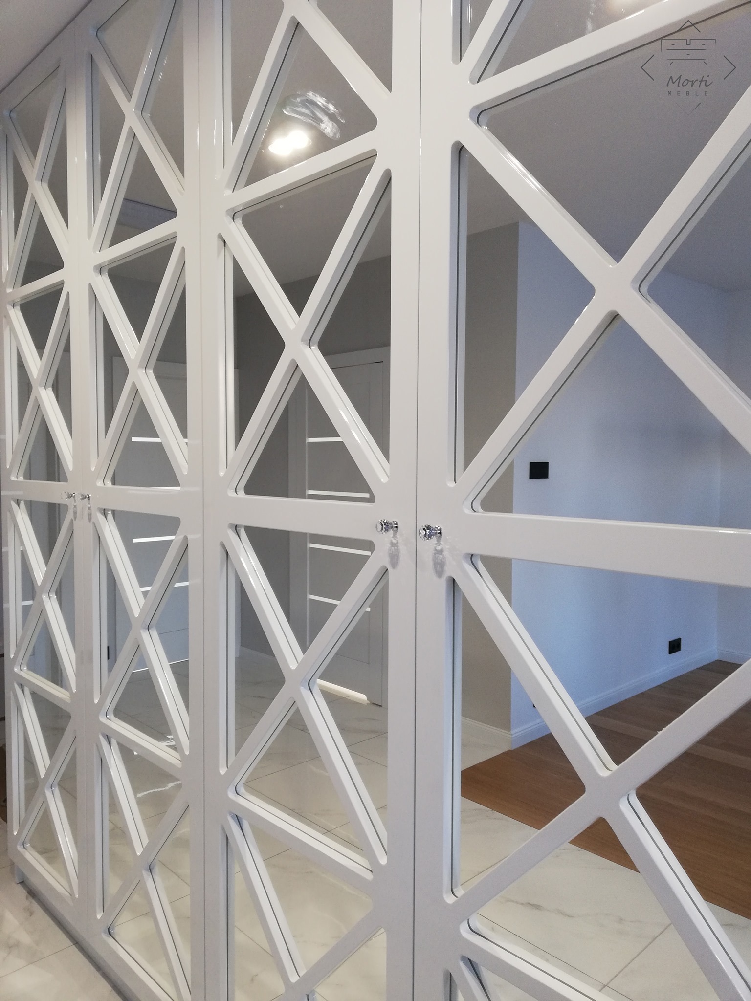 Biała szafa z lustrzanymi drzwiami o geometrycznym wzorze krzyży, kryształowe gałki, widoczny fragment wnętrza z drewnianą podłogą i białymi ścianami.
