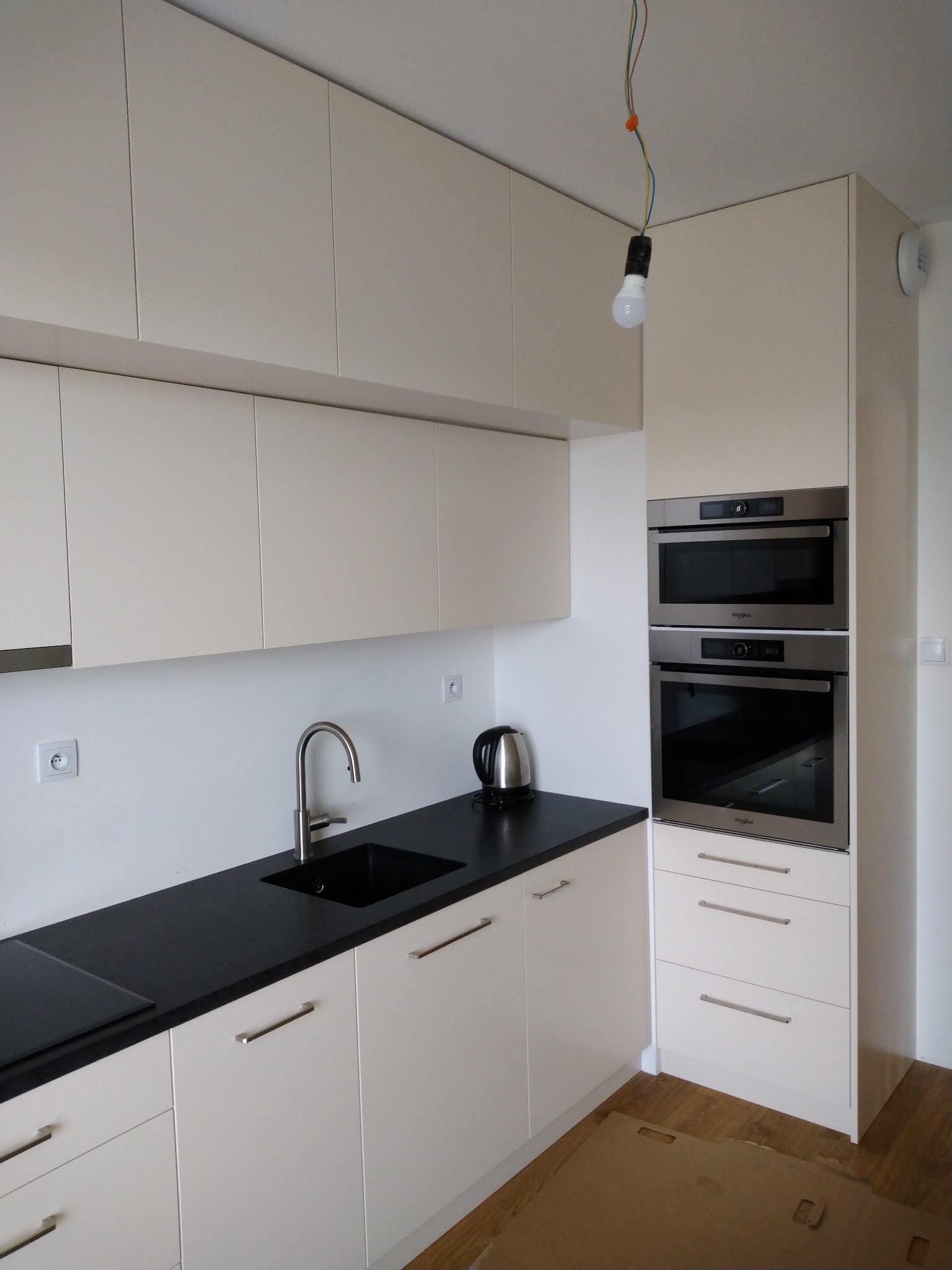 Jasna zabudowa kuchenna z czarnym blatem i wbudowanymi piekarnikami, widoczna wisząca żarówka bez klosza.