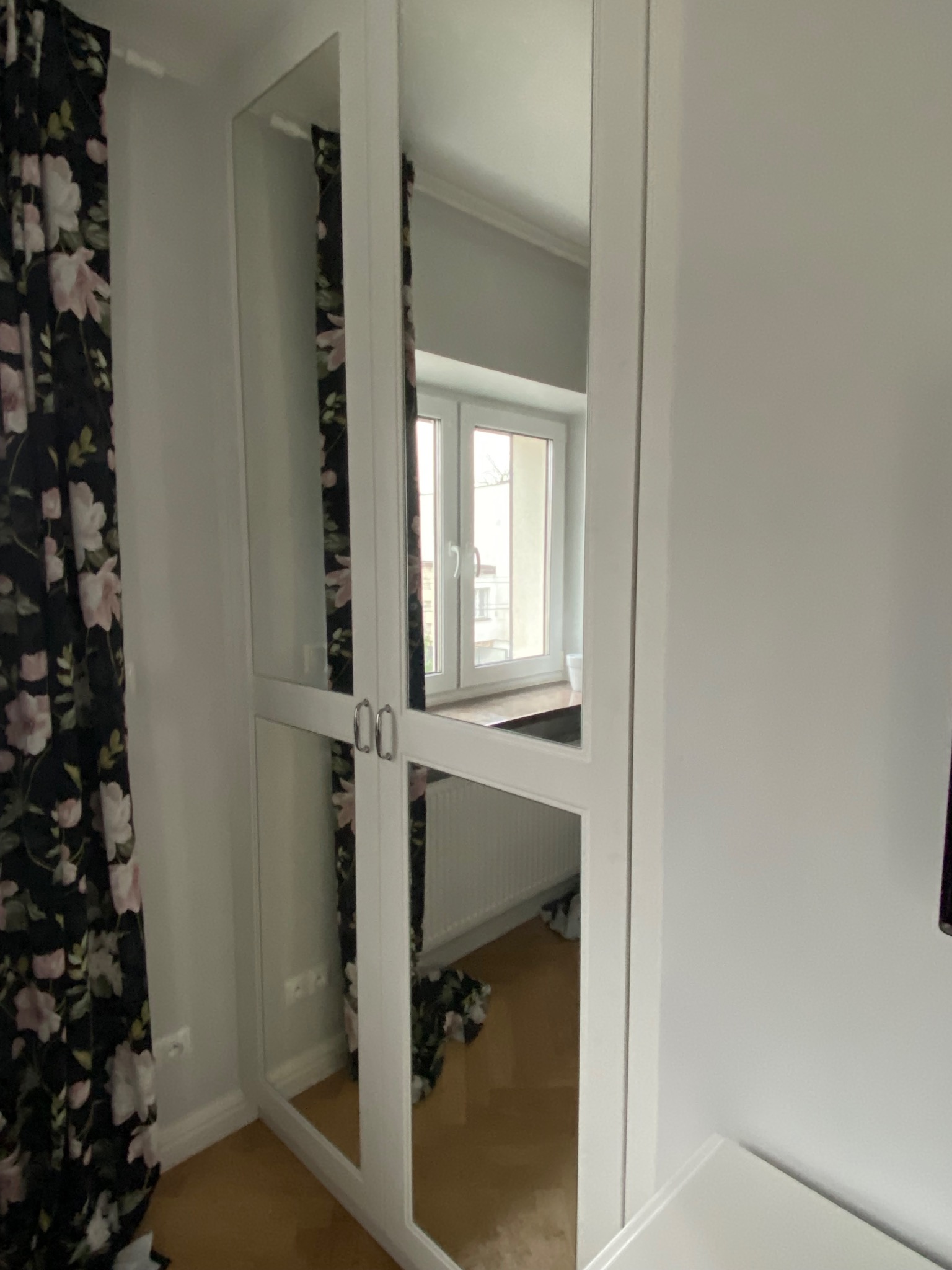 Biała szafa z lustrzanymi drzwiami w pokoju, odbijająca okno i zasłony w kwiaty.