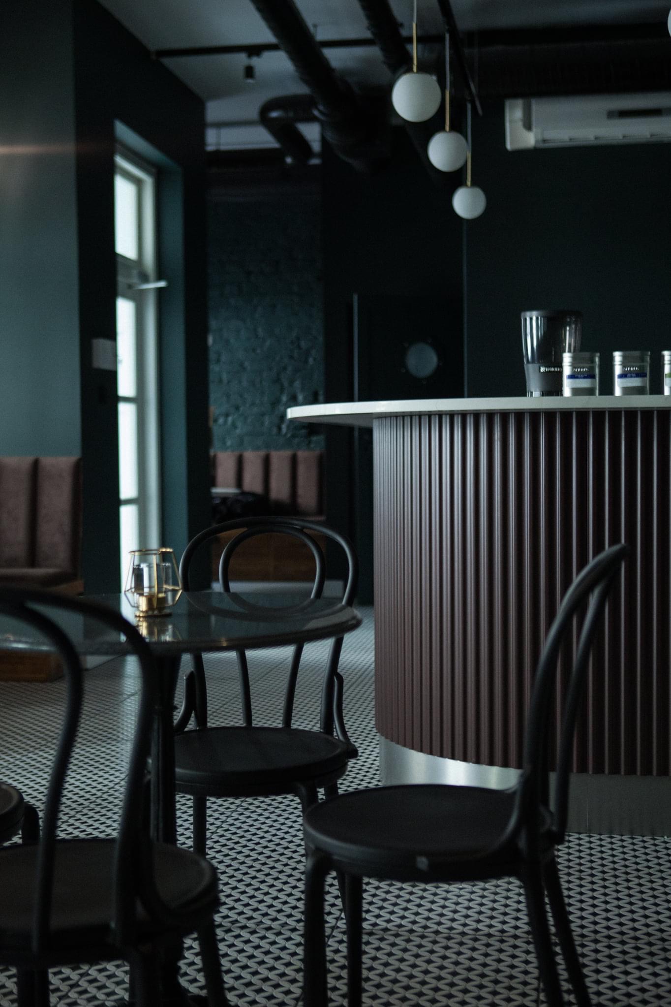 Ciemne wnętrze kawiarni z czarnymi krzesłami Thonet, marmurową ladą barową z ryflowanymi panelami, wzorzystą podłogą i dekoracyjnymi lampami kulami.