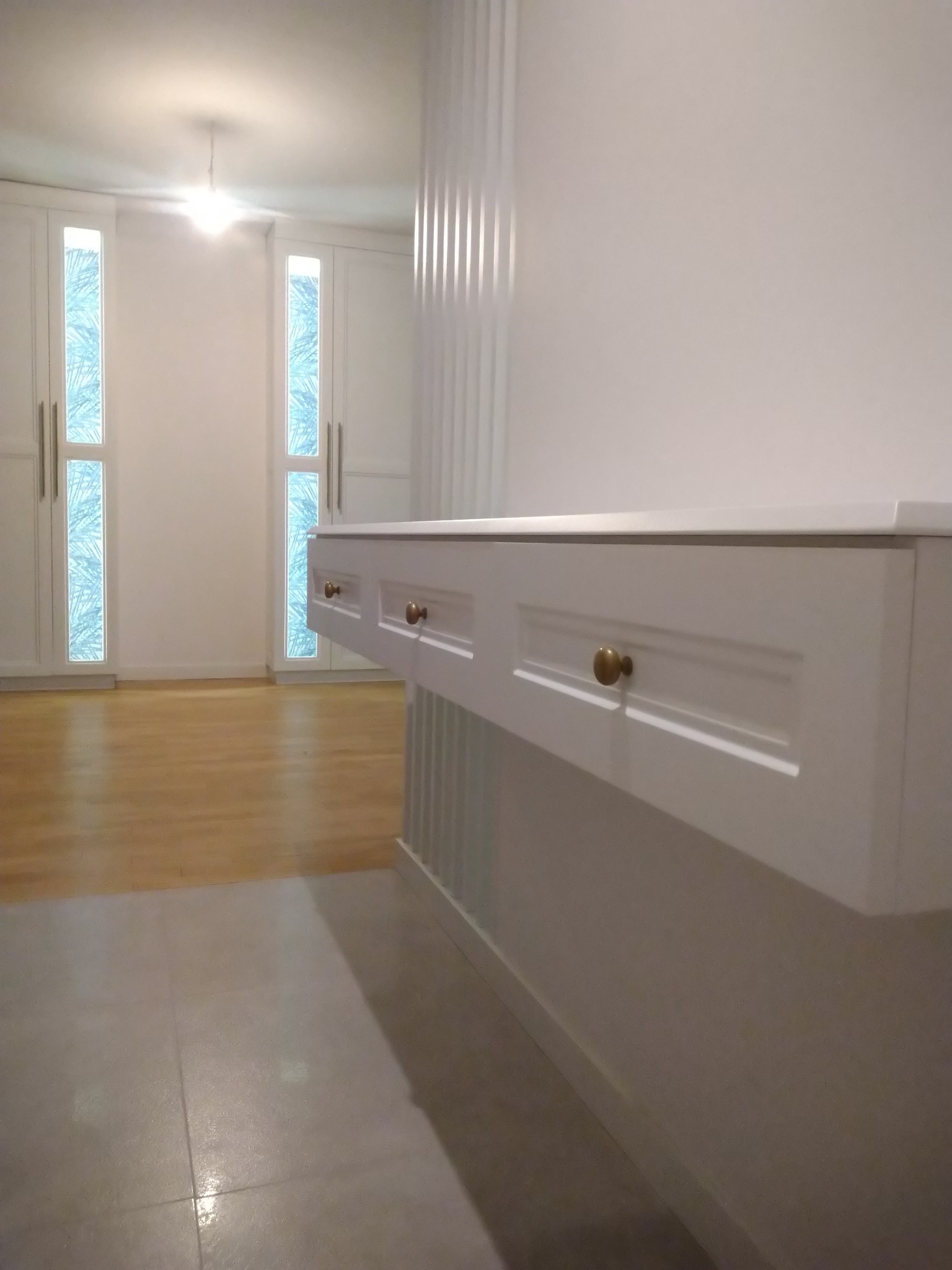 Biała szafka z trzema szufladami i złotymi uchwytami, zamontowana na ścianie w jasnym pomieszczeniu z drewnianą podłogą i białymi szafami w tle.