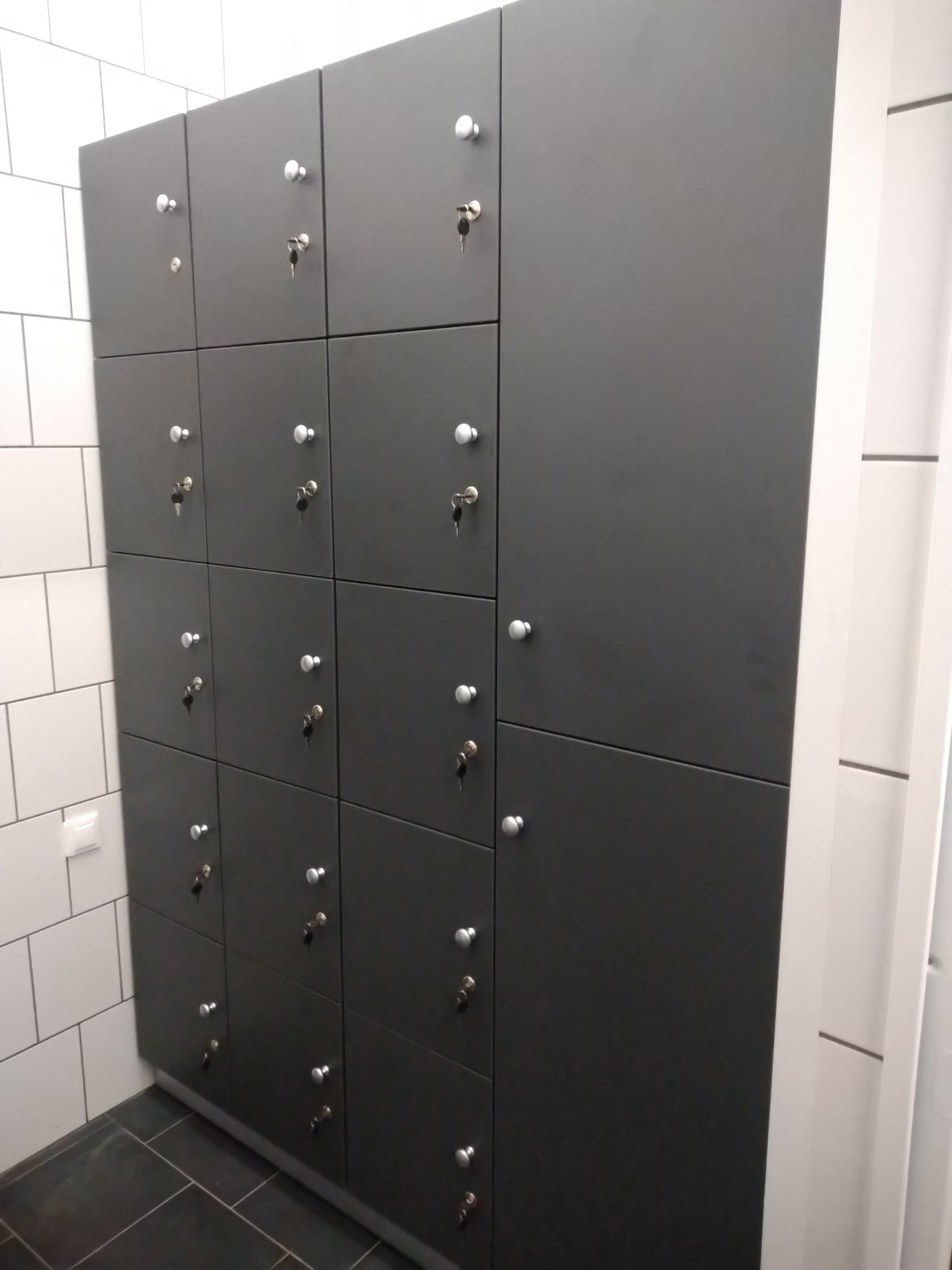Szara zabudowa szafek zamykanych na klucz, białe płytki na ścianie i ciemna podłoga w pomieszczeniu.