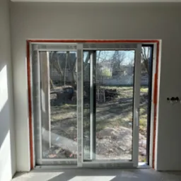 Zamontowane białe drzwi przesuwne z widokiem na ogr&oacute;d, widoczne elementy montażowe i niedokończone wykończenie wnętrza.