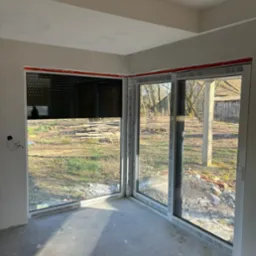 Wnętrze pokoju z narożnym oknem i częściowo opuszczoną roletą zewnętrzną, widok na ogród z elementami gruzu i niedokończonym wykończeniem ścian.