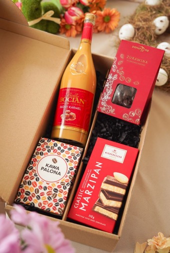 Beżowe pudełko prezentowe wypełnione butelką likieru, kawą, czekoladą marcepanową i żurawiną, udekorowane czarnym wypełniaczem, z kwiatami i pisankami w tle.