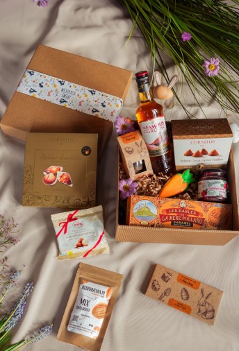 Świąteczny zestaw upominkowy w kartonowym pudełku z dekoracyjną wstążką, zawierający słodycze, alkohol w butelce i drewnianego zajączka, ułożony na kremowym tle z kwiatami.