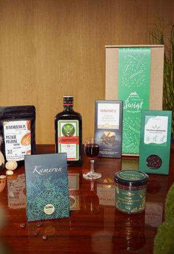 Zestaw prezentowy z pistacjami, likierem ziołowym Jägermeister, czekoladą Hachez, kawą Kamerun, herbatą owocową i kremem orzechowym w słoiku, umieszczony na błyszczącej drewnianej powierzchni.